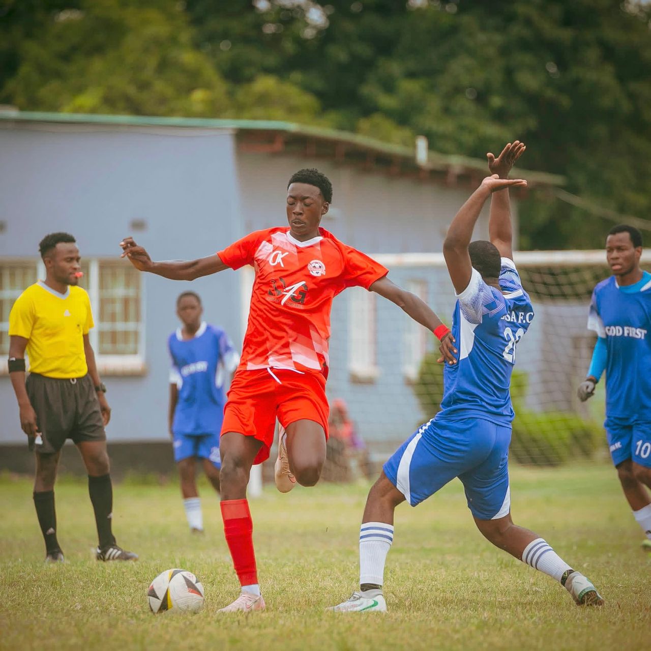 A forward in red shields the ball from a defender in blue during a local league match, the referee watching from behind.